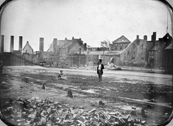 Black-and-white image of rubble in the foreground with a damaged building in the background. A woman with a baby sits in the middle to left of a standing man.¬