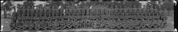 Black-and-white panorama shot of three and a half rows of uniformed soldiers in front of trees and tents. A soldier in the centre of the front row holds a dog on his lap.