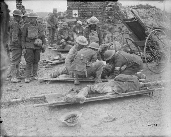 A black-and-white photograph of two men on stretchers. A group of medical personnel is attending to one of the men, while the other man lies on his side. Several soldiers are standing to the left of the stretchers, while others are sitting in the background. The scene is a bombed-out building with only the chimney still standing.