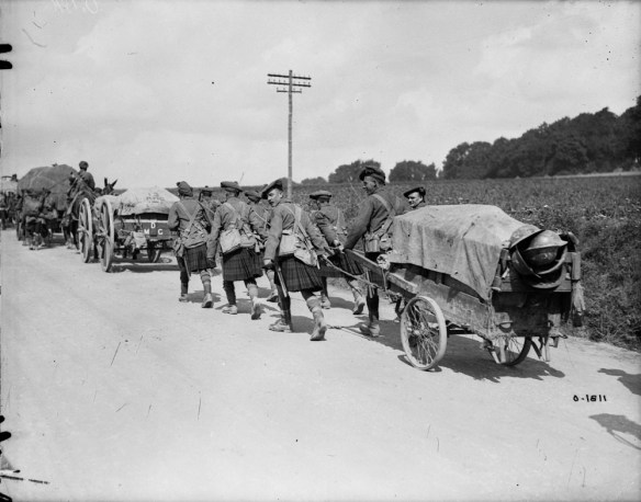 A black-and-white photograph of a convoy of carts moving down the road. A group of Scottish soldiers in full kilt pulls the last cart.