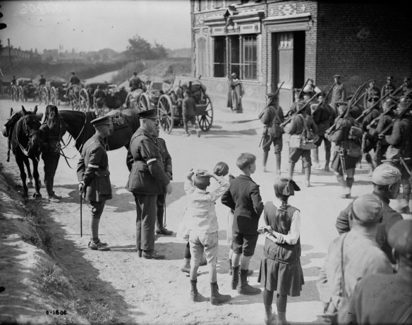 A black-and-white photograph of a column of soldiers marching through a town. Onlookers include some officers as well as children and other civilians.