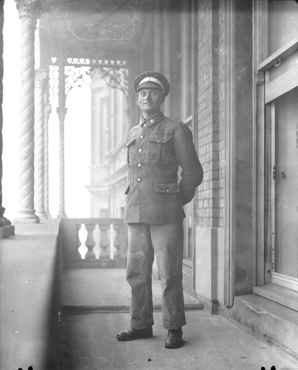 A black-and-white photograph of a young man in uniform standing on a balcony outside. 