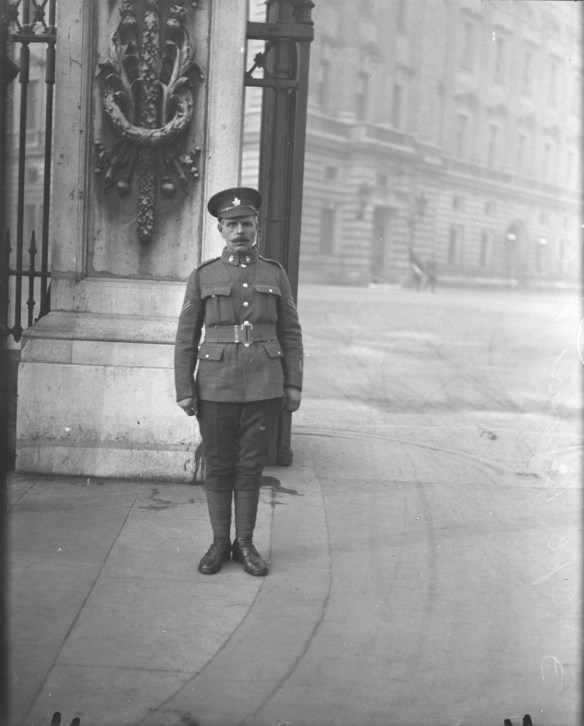 A black-and-white photograph of a soldier wearing a peaked hat adorned with a maple leaf. He is standing at attention in front of a large gate leading into palace grounds. 