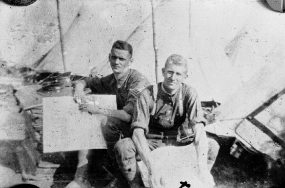 A black-and-white photograph of two young men sitting down in a camp, looking at maps. Behind them can be seen several tents.