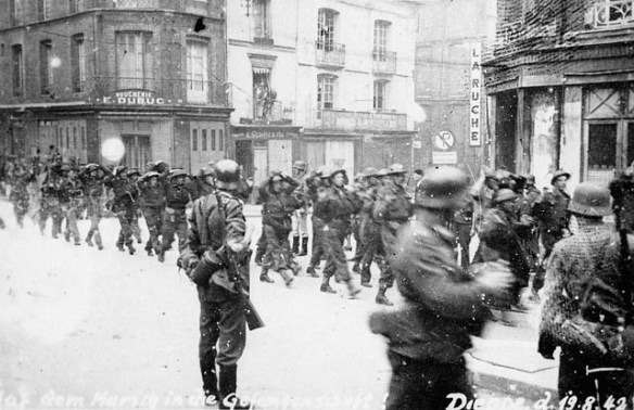 A black-and-white photograph of captured Canadian soldiers being marched in formation through an urban area, with German soldiers guarding them.