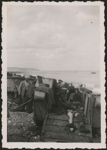A black-and-white photograph of civilians working amid deceased soldiers in a beached landing craft.
