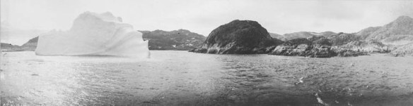 Black-and-white panorama of a large iceberg close to a rocky island shot from a boat. 