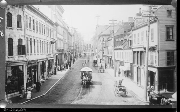 A black-and-white photograph of a horse-drawn streetcar. The rails are located in the centre of the road that has three-story buildings on either side of it. 