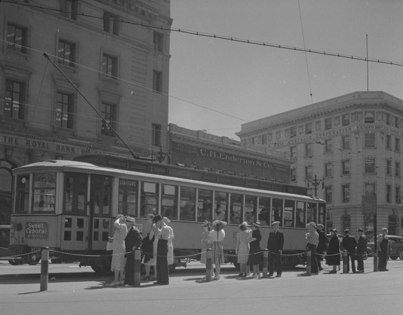 A black-and-white photograph of a lineup of women and men at a pickup location waiting to board an enclosed streetcar.