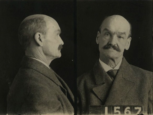A black-and-white mugshot of a slightly balding man with a moustache seen in profile and straight on. He holds up a sign with the number 1567.