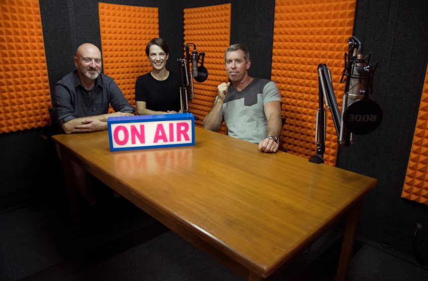 Colour photo of two men and a woman seated in a recording studio with a big sign reading: “On Air”.in front of them.