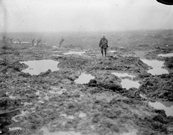 A black-and-white photograph of a soldier walking in a field of mud and puddles.