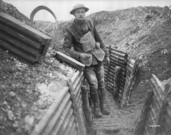 A black-and-white photograph of a muddied soldier leaning on the wall of a trench, smoking a cigarette and looking directly at the photographer.