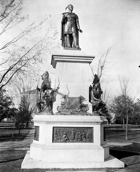 A black-and-white photograph of a bronze statue atop a large stone pedestal decorated with bronze reliefs and statues around the sides. A park with bare trees can be seen in the background.