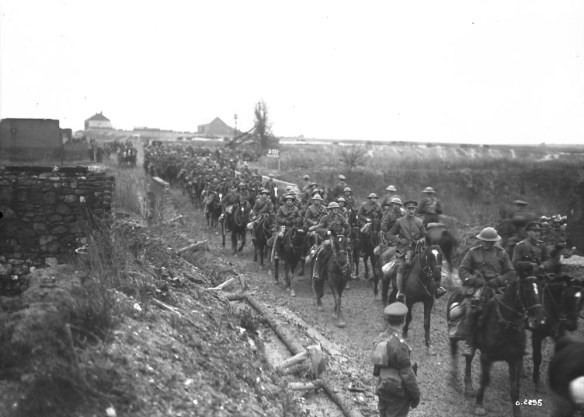 A black-and-white photograph of a line of mounted soldiers riding through a village.
