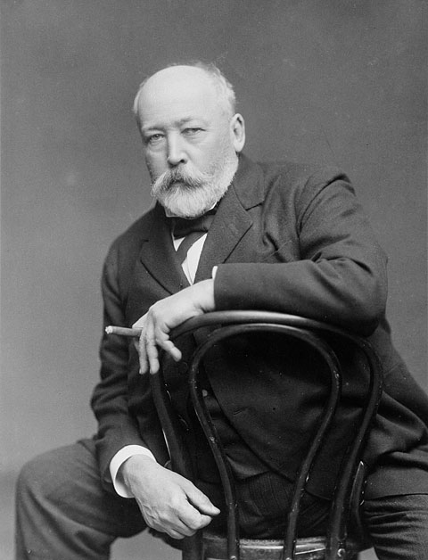 Portrait of a seated man in a suit, straddling a café chair and holding a cigar between two fingers. 