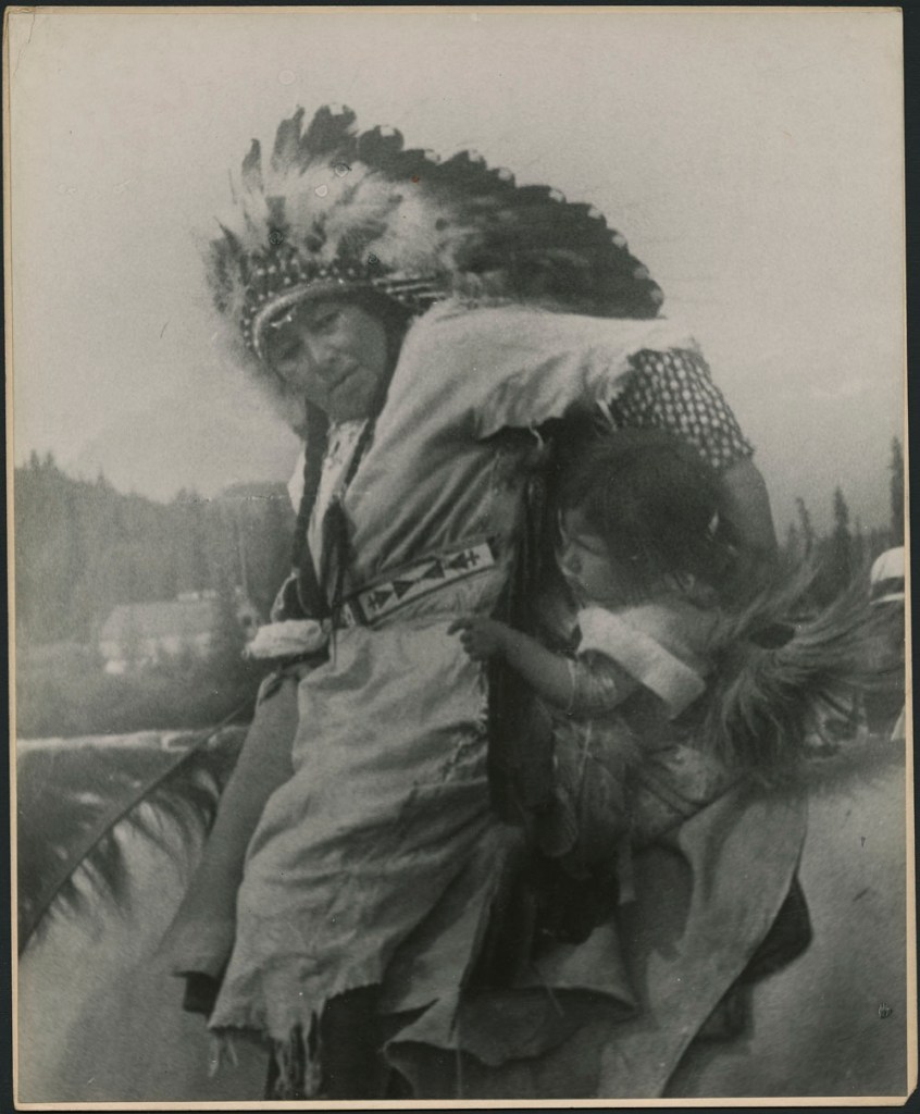 Woman on horseback holding a little girl to her side. The woman is wearing a plain dress with a patterned belt and a feathered headdress over her braided hair.