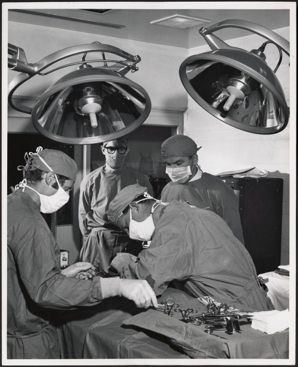 A black-and-white photograph of an operating room scene with four men in surgical gowns and masks gathered around a prone patient who is hidden from view. 