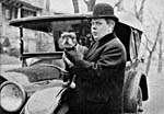 A black-and-white photograph of a man standing in front of a car, holding a small dog and wearing a Bowler hat.