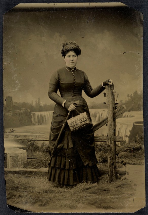 A black-and-white studio portrait of an unidentified woman standing next to a fence with a scene of Niagara Falls in the background.