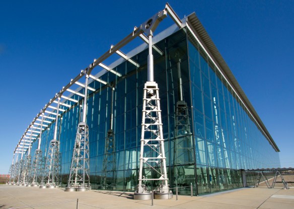 A colour photograph of a large modern building made out of glass with metal pillars.