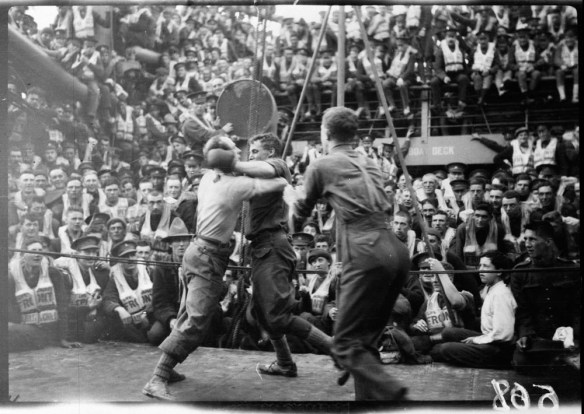 A black-and-white photograph of two boxers fighting on the deck of the SS Justicia, surrounded by the ship’s complement of soldiers. 