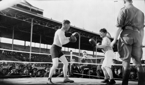 A black-and-white photograph of two soldiers boxing. One wears black trunks and the other wears white trunks. Soldiers outside the ring watch the match. 