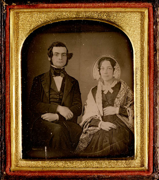 A colour photograph of a sepia-tone image in a wood and gold frame showing a seated man and woman. The man is wearing a suit, waistcoat and cravat. The woman is wearing a bonnet, dress and patterned shawl.