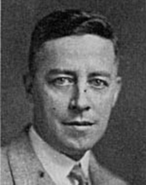 A black-and-white photograph of a man looking right at the camera and wearing a grey suit.