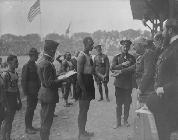A black-and-white photograph of a man dressed in athletic wear, surrounded by other men in similar attire or in uniforms, receiving a medal from an older man in military dress.