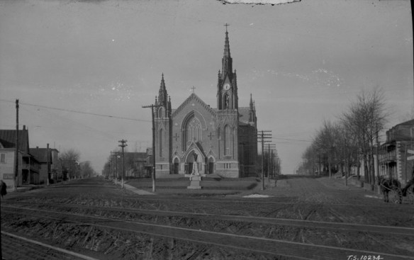 Black-and-white photo of a large church in a small village. Railway tracks can be seen in the foreground.