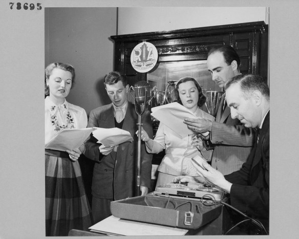 A black-and-white photograph of two women and three men, members of the R. A. Radio Acting Group, reading from a script into a microphone.