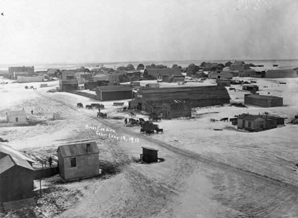 A black-and-white photograph of Cabri, Saskatchewan, from a bird’s-eye perspective. It shows a main dirt road with neighbouring houses and buildings. Some people, horses and wagons gather throughout the town. 