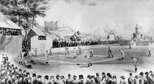 A black-and-white photograph of an outdoor baseball field with a game underway. The crowd watches from the packed stands. The background shows the buildings of the cityscape. 