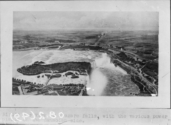 A black-and-white photograph of Niagara Falls from a bird’s-eye perspective. There are various buildings on either side of the border and roads leading up to and alongside the riverbanks.