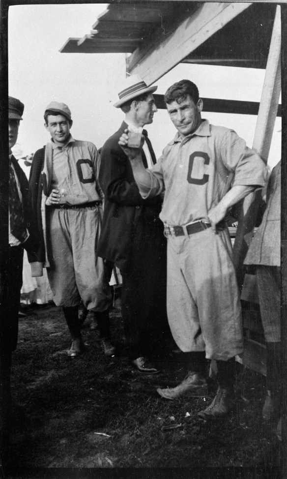 A black-and-white photograph of five men standing around a counter. Two of the men are wearing baseball uniforms with a large letter ‘C’ on the chest. The other men are wearing suits and hats. One of the uniformed men is holding up a drink and looking towards the camera.
