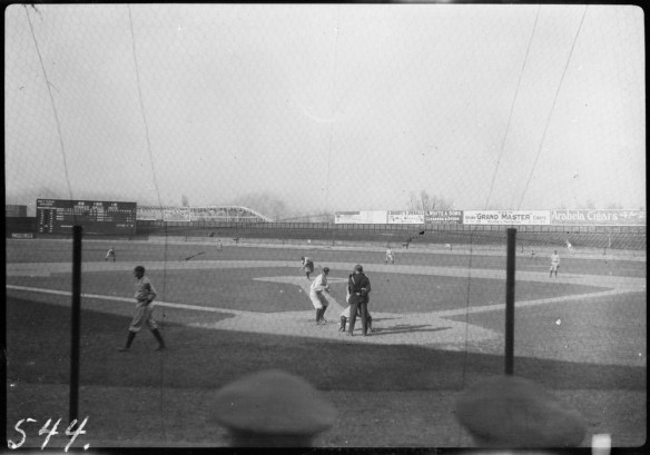 A black-and-white photograph of a baseball game from behind home plate. A player is at the plate as a pitch comes in. The umpire stands behind him to make the call.