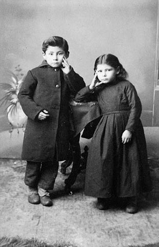 A black-and-white studio photograph of two children leaning against a side table, each with a hand on a cheek.