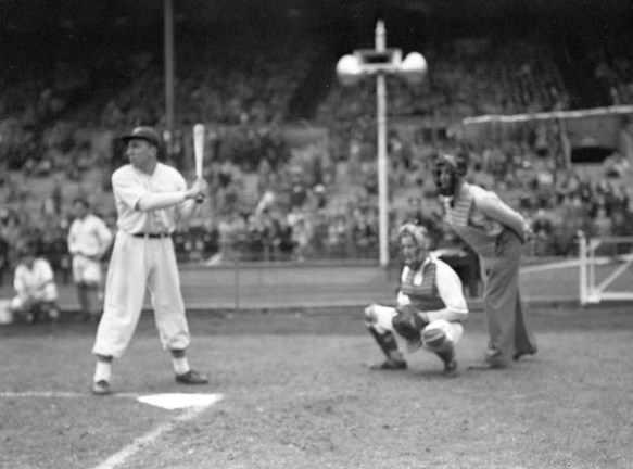 A black-and-white photograph of a baseball game. A player stands with a bat and behind him are a catcher and an umpire. In the background are players watching the play and spectators in the stands.
