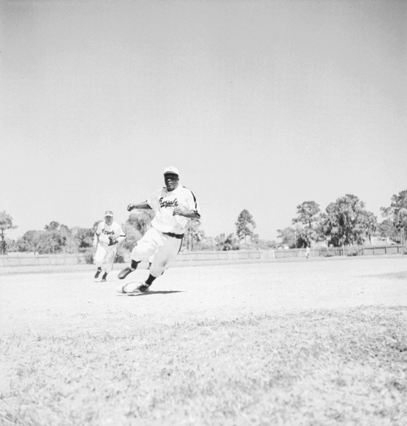 A black-and-white photograph of a baseball player rounding the bases as a player on the opposing team tries to catch up to him. 