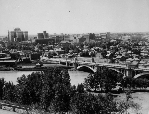 A black-and-white photograph of Calgary, Alberta, from a bird’s-eye perspective. The Bow River and a bridge are in the foreground with a number of homes and larger buildings in the background.