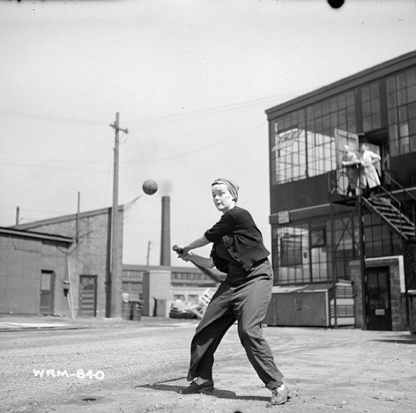 A black-and-white photograph of a woman in work clothes and a headscarf swinging a baseball bat at a ball. She stands in a vacant lot with industrial buildings and other structures in the background.