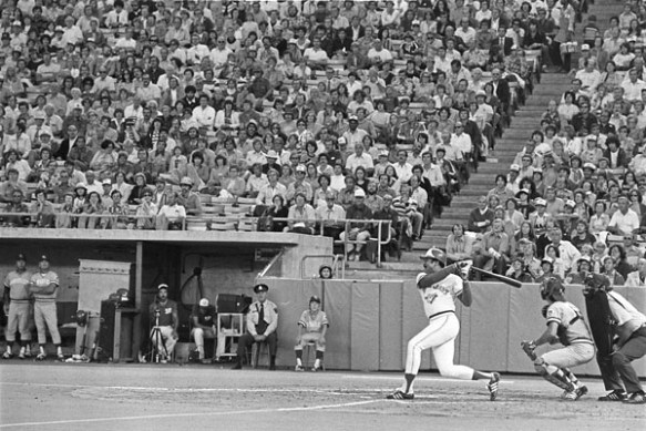 A black-and-white photograph of a baseball game. There’s a man who has just swung at the ball. Behind him is a man wearing catcher equipment and crouching, while behind him is an umpire, also crouching. In the background are players in baseball uniforms and a man wearing police uniform, all watching the action. Behind them spectators are seated in the stands.