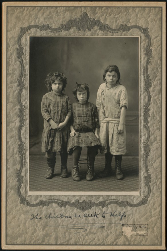 A black-and-white studio photograph of three children. One is sitting in a chair and the two others stand beside.