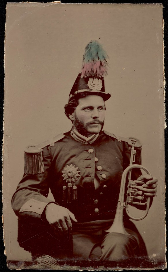 A black-and-white photograph of a man sitting, wearing a band uniform and cap with feathers. The man is holding a flugelhorn resting on his left knee. 