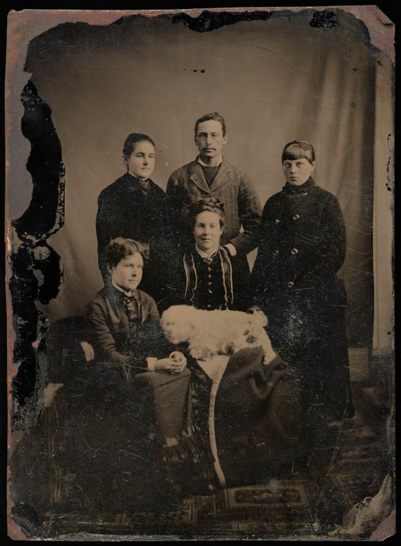 A black-and-white photograph of a mother, three daughters, a son and a dog posing for a family portrait. 