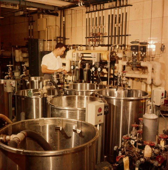 A colour photograph of a man in a chemical mixing laboratory preparing various fluids used in developing processes at the National Film Board of Canada.