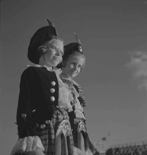 A black-and-white photograph of two girls who are standing and wearing tams, matching jackets and kilts. 
