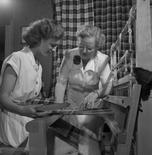 A black-and-white photograph of two women at a loom. The woman sitting on the left holds a shuttle. The woman standing on the right inspects the tartan pattern and weave. 