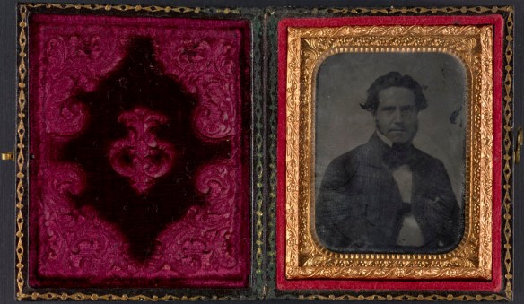 A colour image of a paper case with red velvet. On the right-hand side, a black-and-white photograph in a brass mat of a man wearing a dark jacket.